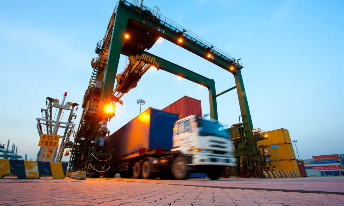 Container terminal at dusk: a gantry crane and truck moving shipping containers — ocean freight and inland delivery when importing inflatables from China